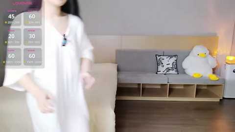 Video of a young Asian woman in a white robe, standing in a modern living room with a light grey sofa, white pillow, and plush toy duck.