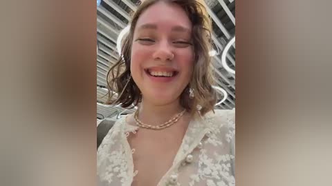 Video of a smiling woman with short, wavy brown hair, wearing a sheer, floral-patterned blouse and a beaded necklace. She stands in a modern, glass-walled building with a metal framework in the background.