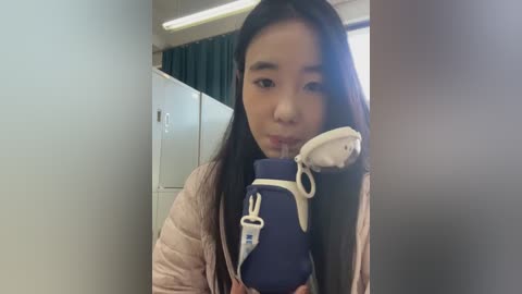 Video of a young Asian woman with long black hair, wearing a light jacket, holding a blue medical bag with a white cross emblem, standing in a dimly lit, modern office with white walls and green curtains.