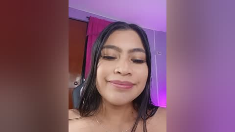 A close-up video of a smiling Latina woman with long black hair, wearing makeup, in a room with purple lighting and red curtains in the background.