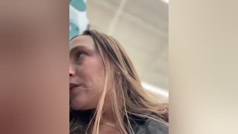 A video of a blonde woman with wet hair, captured from a side angle, showing her profile and shoulder. She is wearing a dark jacket, and the background features a blurred, beige wall and a white ceiling.