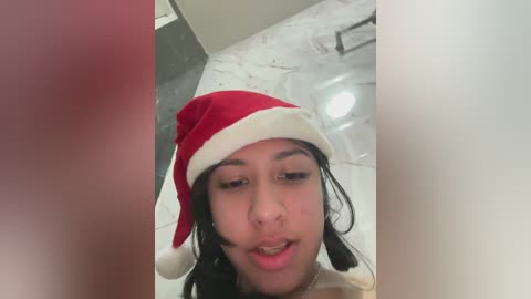 Video of a young woman with light brown skin and long black hair, wearing a red and white Santa hat, standing in a modern bathroom with marble flooring and a clear shower door.