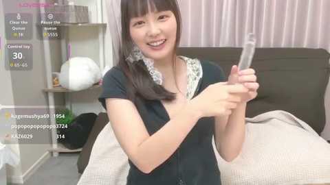 A video of an East Asian woman with long black hair and fair skin, wearing a black shirt, holding a syringe, smiling in a bedroom with a bed and white curtains.