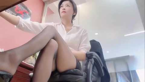Video of a Chinese woman with short black hair, wearing a white shirt, black shorts, and sheer black stockings, seated on a black chair in a modern office with red walls and a framed abstract painting.