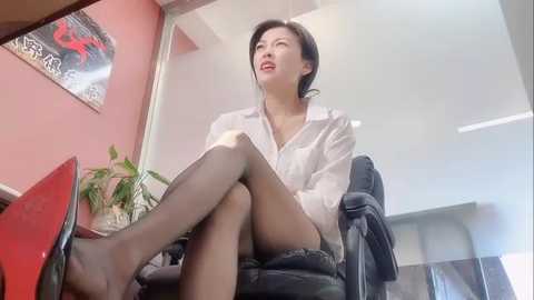 Video of an Asian woman with short black hair, wearing a white blouse and black stockings, sitting on a black office chair, legs crossed, in a modern office with red walls and a plant.
