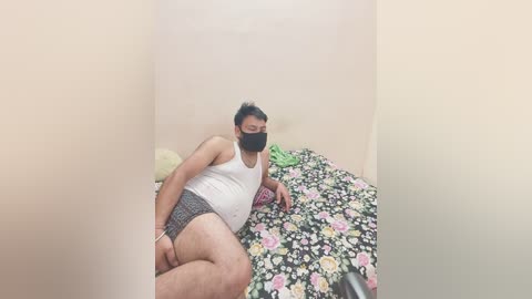 A video shows a middle-aged, overweight man with dark hair and a beard, lying on a floral-patterned bed in a small, plain room, wearing a white tank top and black briefs.