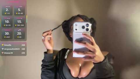 A video of a woman with medium skin tone, wearing a black bra that accentuates her large breasts, taking a selfie in a mirror. The background is blurry, showing a bathroom with a beige wall.