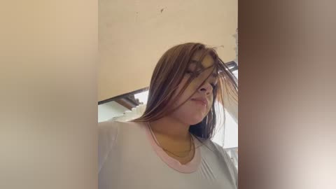 A video of a young woman with light skin and long, straight brown hair, wearing a white shirt and a gold necklace, standing against a beige wall with a window in the background.