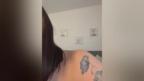 A video of a woman's back, showing a large blue floral tattoo on her right shoulder blade. The room has a minimalist aesthetic with white walls and abstract art.