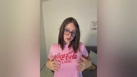 A video of a young woman with fair skin, straight brown hair, and glasses, wearing a pink \"New Classic\" T-shirt, standing in a minimalist room with white walls and a gray couch.