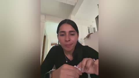 A video of a young woman with long black hair, fair skin, and a neutral expression, wearing a black top, in a dimly lit, modern kitchen with white cabinets and a microwave.