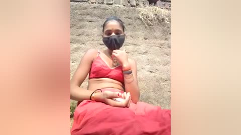 Video of an Indian woman with a medium complexion, wearing a red saree, black mask, and a bracelet, sitting on a dirt path, surrounded by dry grass and a stone wall.