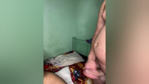 Video of a nude man in a dimly lit, mint-green room. He is standing next to a bed with colorful, patterned sheets, holding his penis. The background includes a wooden headboard and a messy, unmade bed.
