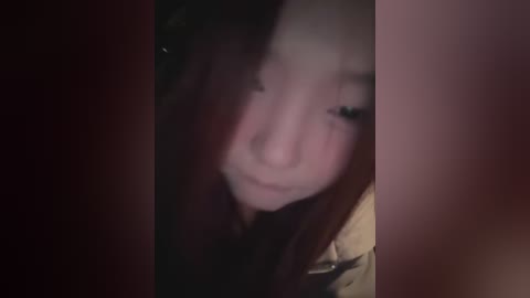 A dimly-lit, grainy video of a young Asian woman with long dark hair partially obscuring her face, wearing a beige jacket, with a blurred background.