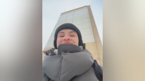 A video of a person wearing a black beanie, gray puffer jacket, and black gloves, standing in front of a tall, beige, rectangular building. The image is taken from a low angle, emphasizing the person's upward gaze.