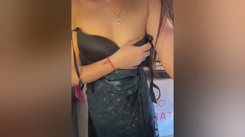Video of a woman with fair skin, wearing a black strapless dress, holding the fabric at her chest, revealing a tattoo on her left forearm. She has long, straight brown hair and a silver necklace with a pendant.