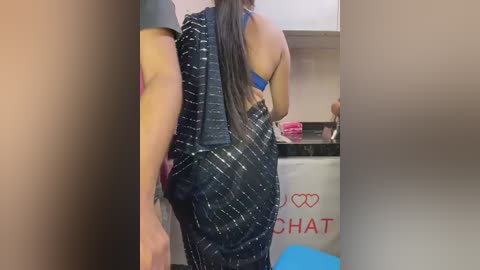 Video of a woman with long dark hair, wearing a blue bra and a black patterned dress, walking away from the camera in a laundry room with white cabinets and a \"CHAT\" logo.