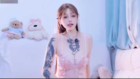Video of a slender East Asian woman with light skin, long brown hair, and intricate tattoos on her chest and arms, wearing a pink lace lingerie set. She sits on a white bed with stuffed animals and light blue curtains in the background.