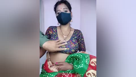 Video of a South Asian woman with medium skin tone, wearing a colorful floral blouse, a green and red sari, a black face mask, and red nail polish.