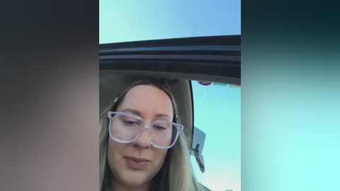 Video of a young woman with fair skin and light brown hair, wearing clear glasses and a neutral expression, sitting in a car with a light blue sky visible through the window.