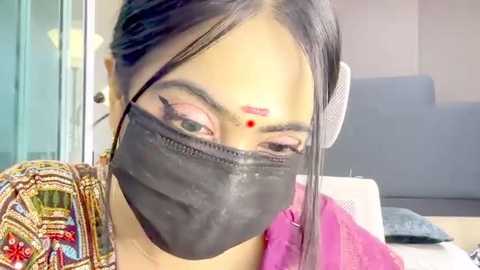 Video of a woman with light brown skin, wearing a black mask, pink headband, and colorful blouse, sitting indoors next to a sofa.