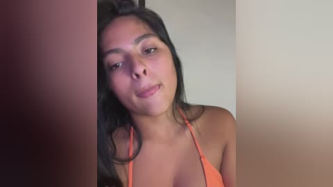 Video of a young, light-skinned woman with long black hair, wearing an orange bikini top, smiling slightly, partially obscured by a vertical object on the left. Background is a plain, light-colored wall.