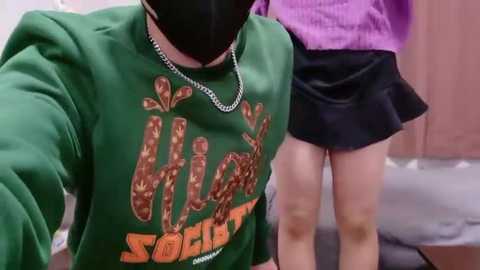 A video showing a person in a green \"Society\" sweatshirt with orange lettering, black face mask, and silver chain necklace, standing beside a person in a purple sweater and black skirt.