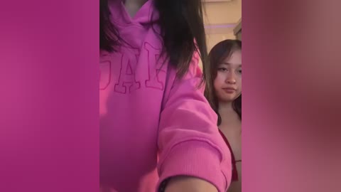 Video of two Asian women; one in a pink hoodie with \"JACK\" text, the other in a black top. They are indoors, with pink lighting.