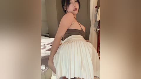 Video of an Asian woman in a brown tank top and white pleated skirt, posing seductively in a dimly lit bedroom with a bed, door, and wall.