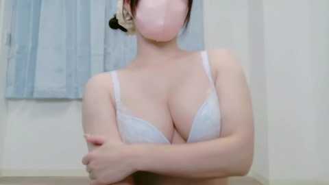 Video of an Asian woman with fair skin, wearing a white lace bra, crossed arms, and a headset, standing in a simple room with white walls and light blue curtains.