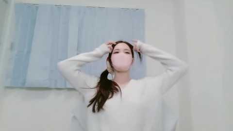 Video of a young Asian woman in a white long-sleeve shirt, adjusting her ponytail in a brightly lit, minimalistic room with light blue curtains.