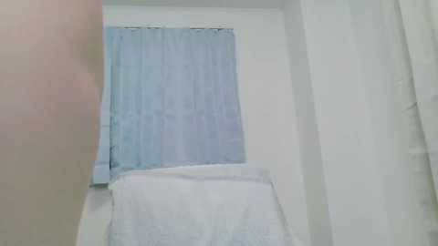 Video of a small, white-walled room with a light blue curtain covering a window. A neatly made bed with white sheets is visible in the foreground, creating a clean and minimalistic ambiance.