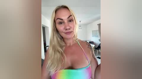 Video of a blonde woman with long hair, wearing a colorful, low-cut bikini top, standing in a modern, bright living room.