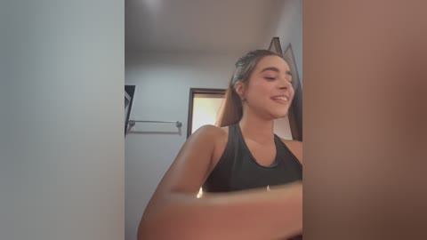 Video of a young woman with fair skin, light brown hair, and a medium build, wearing a black sports bra. She's smiling, standing in a dimly lit bathroom with a towel rack and wooden door in the background.