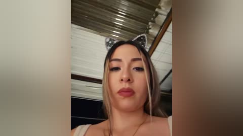 Video of a young woman with light brown skin, wearing a white top and cat ears, indoors with a corrugated metal ceiling.