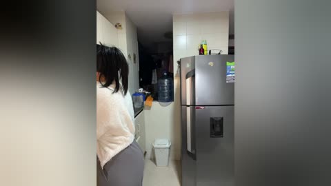 A video shows a woman in a white shirt and gray pants standing near a kitchen counter with a fridge and water dispenser, a sink, and a white trash can.