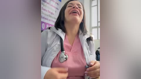 Video of a cheerful woman in a pink scrub top, white jacket, and stethoscope, smiling broadly in a medical office with white walls and a purple informational poster.