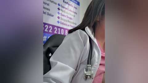 A video of a person with long black hair, wearing a light gray jacket, sitting in a chair. A stethoscope is around their neck. The background shows a partially blurred wall with a sign listing phone numbers and services.