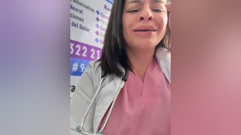 Video of a smiling Latina woman with medium skin tone, long dark hair, wearing a pink lab coat, standing against a purple and blue informational poster in a medical setting.