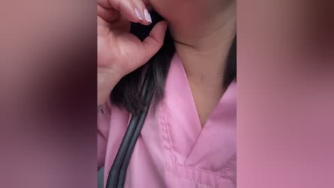 Video of a person's neck and shoulder, wearing a pink medical scrub top with a black stethoscope around their neck. The focus is on the hand holding the stethoscope, with a manicured nail visible.