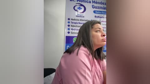 A video shows a middle-aged woman with shoulder-length dark hair, wearing a pink hospital gown, sitting contemplatively in a medical office with a poster featuring Dr. Monica Mendez Guzm\u00e1n.