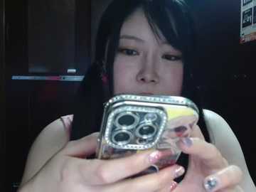 A video shows an East Asian woman with long black hair, wearing a sleeveless top, holding a silver iPhone with a yellow case, looking intently at the screen. The background is dark, and the lighting is dim.