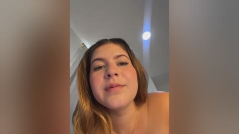 A candid video of a fair-skinned young woman with light brown hair, wearing minimal makeup, and a subtle smile, taken indoors with a blurred background.