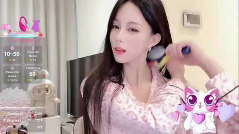 A video of a fair-skinned East Asian woman with long black hair, applying makeup with a brush, wearing a pink floral shirt. Background shows a bed, curtains, and a virtual heart overlay.