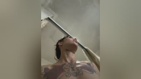 Video of a tattooed woman with dark hair, topless, gazing upward, showcasing detailed tattoos on her chest and neck. The background features a ceiling and a metal shower rod.