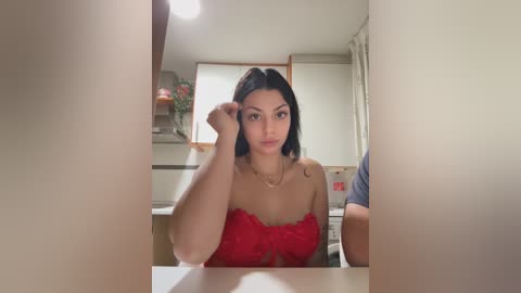Video of a young woman with long black hair, wearing a red lace top, sitting at a table in a kitchen with white walls and a small window.