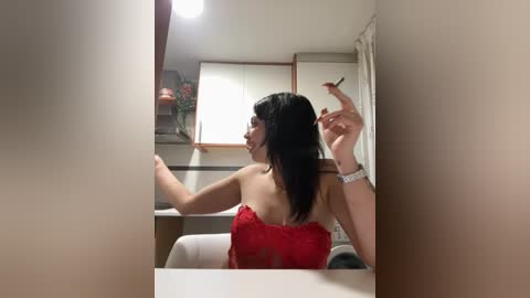 A video of a young woman with long black hair, wearing a red lace top, smoking a cigarette, sitting in a dimly lit room with beige walls and a white table.