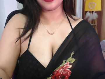 A close-up video of a light-skinned Asian woman with long black hair, wearing a black halter top revealing cleavage, and a red rose. The background features a yellow lamp and white wall.