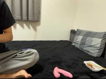 Video of a person kneeling on a black bedspread, wearing a black shirt and gray pants, with a pink sock beside them, in a room with gray curtains and a white wall.