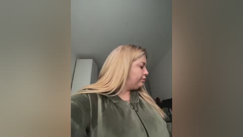 A video of a fair-skinned, blonde woman with long hair, wearing a green jacket, standing in a dimly lit, beige-walled room.